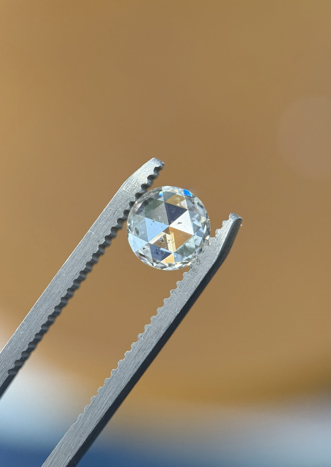 Close-up of 1.04CT round rose cut diamond held in gemstone tweezers against a neutral background, highlighting the brilliance and facet detail of this F color I1 clarity stone from the Gahcho Kué Mine, Canada.