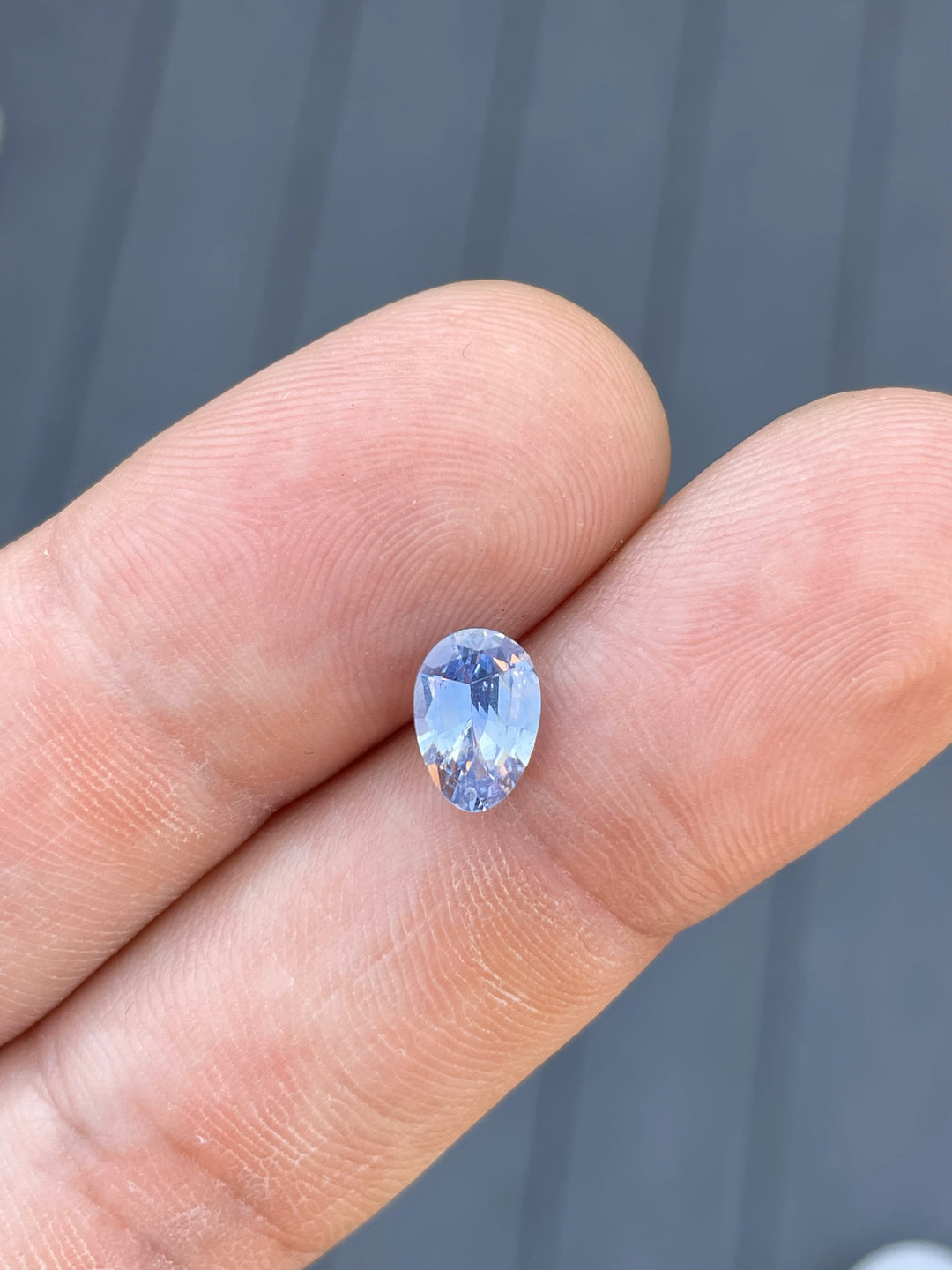 A 1.74-carat sapphire in a custom Poval cut rests on the fingertip. The stone showcases a delicate Ceylon denim blue color, highlighting its pristine facets and oval shape. The dimensions of the untreated sapphire are 9.18mm in length, 6.38mm in width, and 3.82mm in height, originating from Sri Lanka.