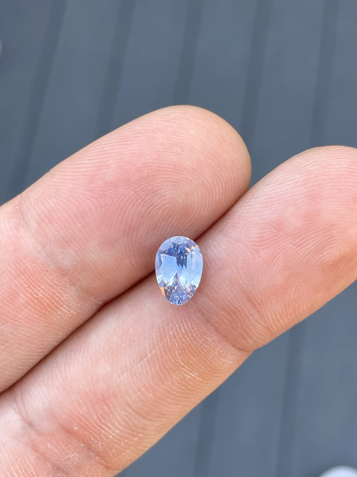 A 1.74-carat sapphire in a custom Poval cut rests on the fingertip. The stone showcases a delicate Ceylon denim blue color, highlighting its pristine facets and oval shape. The dimensions of the untreated sapphire are 9.18mm in length, 6.38mm in width, and 3.82mm in height, originating from Sri Lanka.