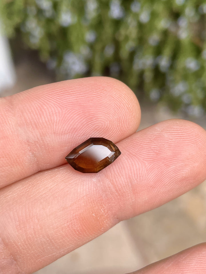 A 3.78-carat earthy brown cognac sapphire sits on a fingertip, highlighting its custom leaf portrait cut and smooth surface. The sapphire from the Umba River, Tanzania, is untreated and measures 12.76mm by 4.78mm by 3.80mm.