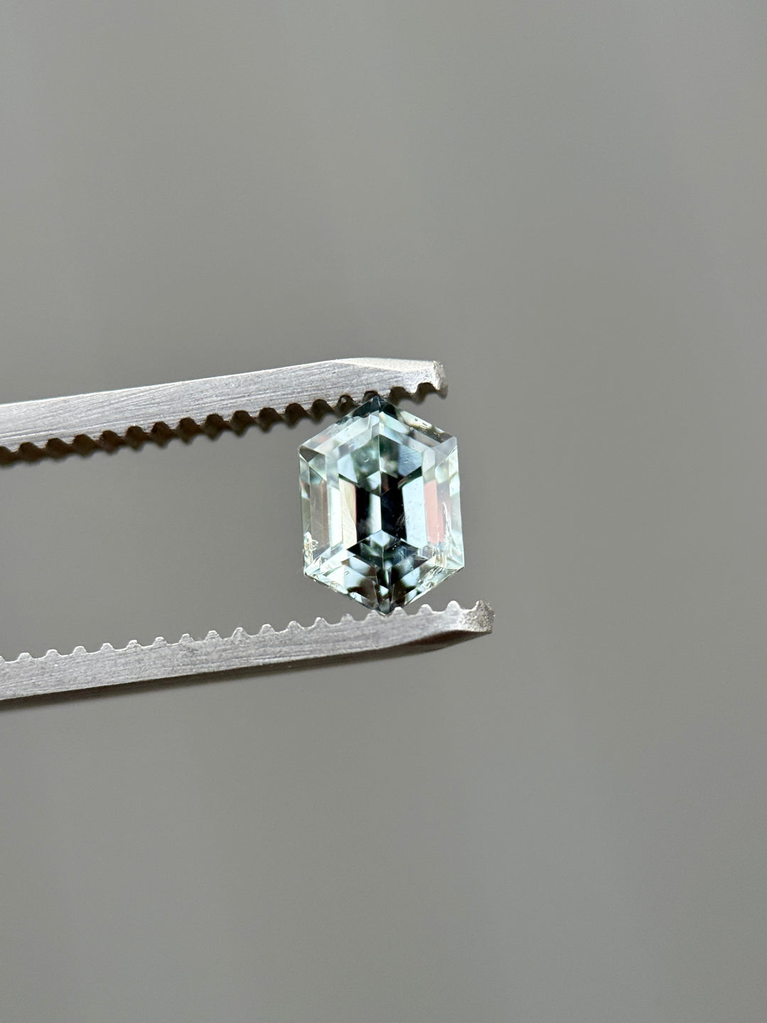 Color-shift Montana sapphire held in jeweler’s tweezers, showing light seafoam to grey silver tones in a 1.22 carat precision elongated hexagon step cut, untreated and ethically sourced from Montana, USA.