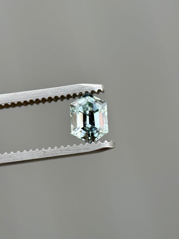 Color-shift Montana sapphire held in jeweler’s tweezers, showing light seafoam to grey silver tones in a 1.22 carat precision elongated hexagon step cut, untreated and ethically sourced from Montana, USA.