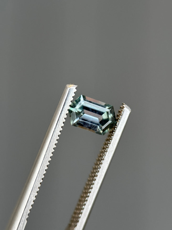 Color shift Montana sapphire held in precision tweezers, showing elongated hexagon step cut facets with light seafoam tones shifting toward soft purple under studio lighting