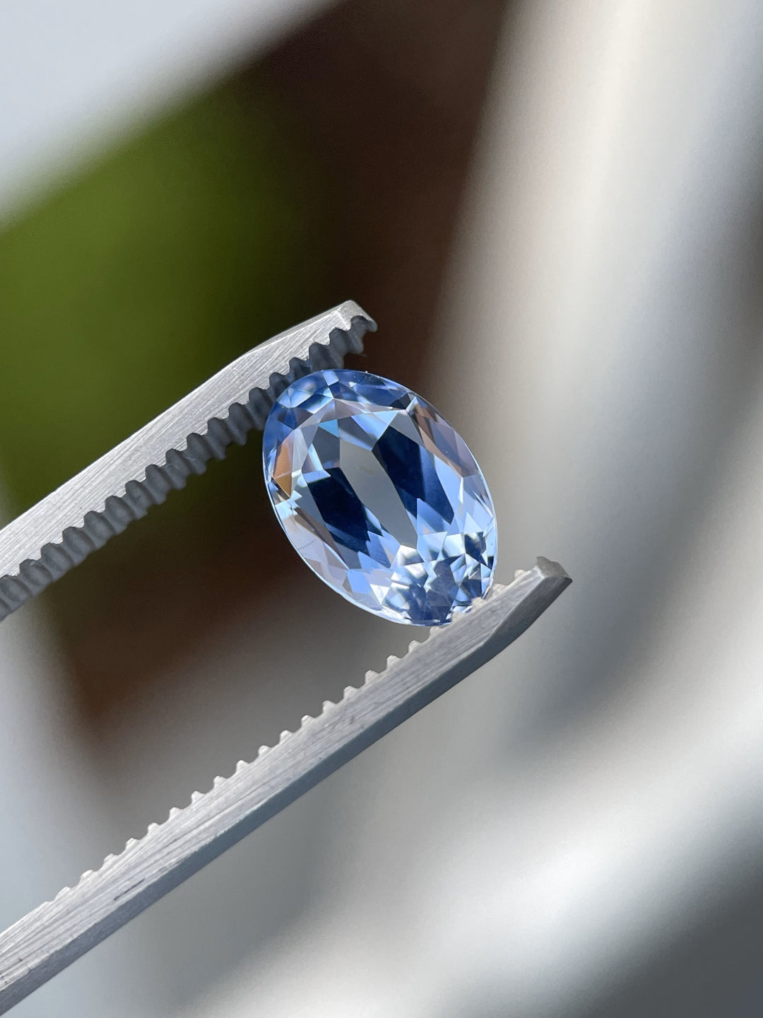 Cornflower blue sapphire, 2.50 carats, securely held in tweezers, highlighting its mix-cut oval facets and vibrant blue hue against soft natural light.
