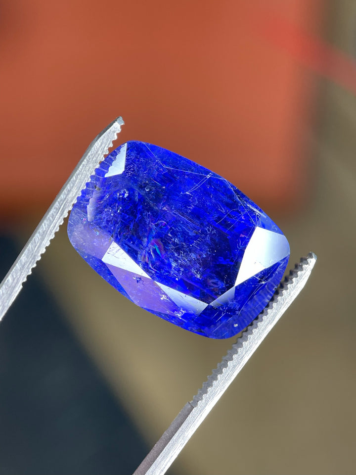 A close-up of a 27.31-carat tanzanite held by gem tweezers, highlighting its cushion shape, deep saturation, and natural internal characteristics.