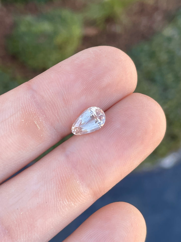 2.45 carat pastel champagne peach sapphire, custom brilliant teardrop cut, showcased against fingertips for size reference.