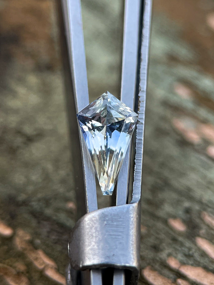 A custom mix kite-cut sapphire with a striking yellow and cornflower blue parti color held by tweezers against a textured natural background. The sapphire has a sharp, geometric cut with visible brilliance and precision.