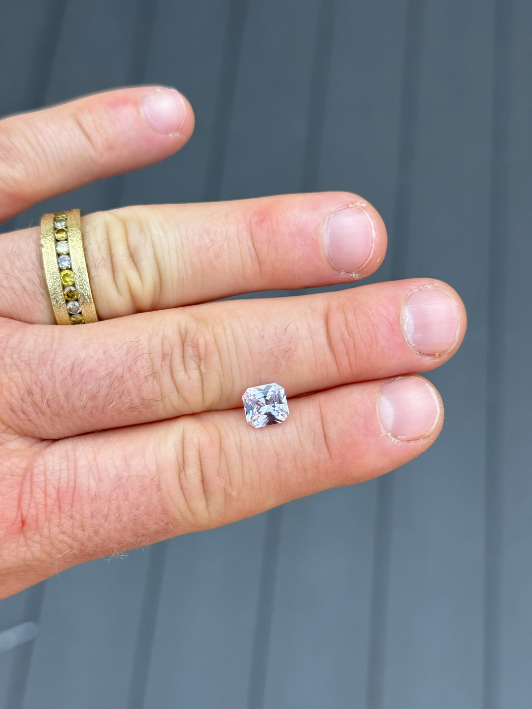 Hand shot of a 2.14-carat natural lavender sapphire resting on the finger for scale, showcasing its radiant octagonal cut with a soft purple-pink hue, against a muted gray panel wall.