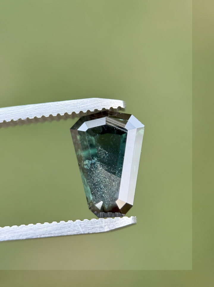 A rich 3.65 carat Kenyan sapphire in a custom step-cut shield shape with deep blue, green, and peacock tones, held securely by gem tweezers against a soft, neutral-toned blurred background.