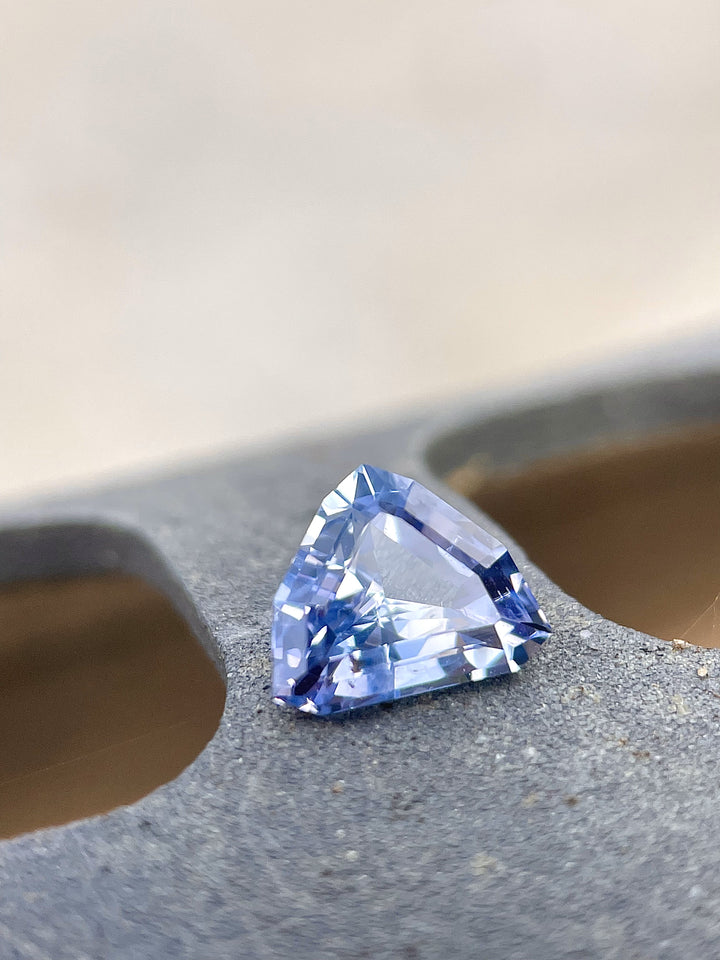 A 1.77-carat lavender and powder blue sapphire sits on a textured surface, highlighting its custom mix shield cut. The gemstone's natural color shift and clean facets are emphasized, with dimensions of 6.99mm by 8.73mm by 4.03mm. Sourced from Sri Lanka and untreated.