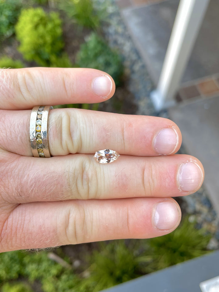 A 2.07-carat peach and champagne sapphire with a custom geometric shield brilliant cut, displayed on a fingertip for scale. The gem's brilliant facets and warm tones are evident. The stone, sourced from Sri Lanka, measures 11.02mm by 5.87mm by 3.94mm and is untreated.