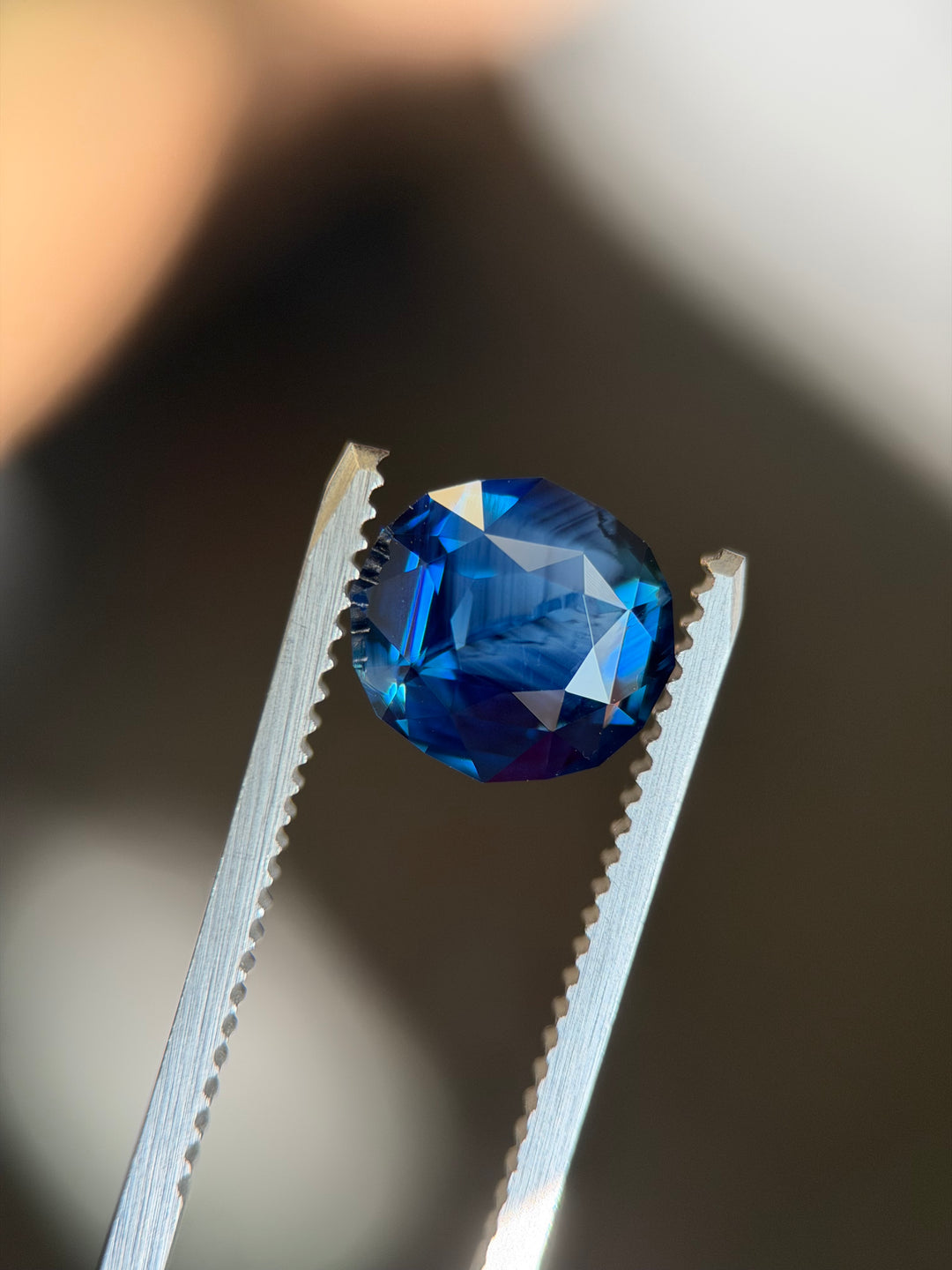 Custom geo oval precision cut royal blue opalescent sapphire with natural growth patterns held in gem tweezers under natural light.