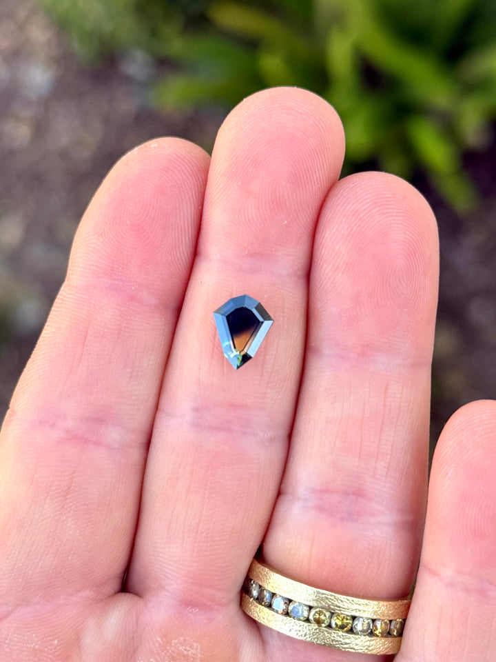 Custom shield portrait cut sapphire resting on fingertips, showing transparent yellow and blue-green color zones with geometric symmetry.