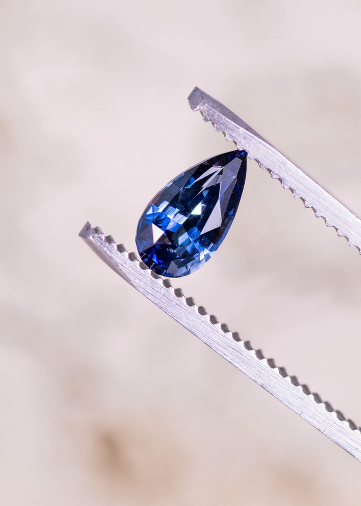 Loose denim blue sapphire from Madagascar held in tweezers displaying the mixed brilliant pear cut and rich blue tone.