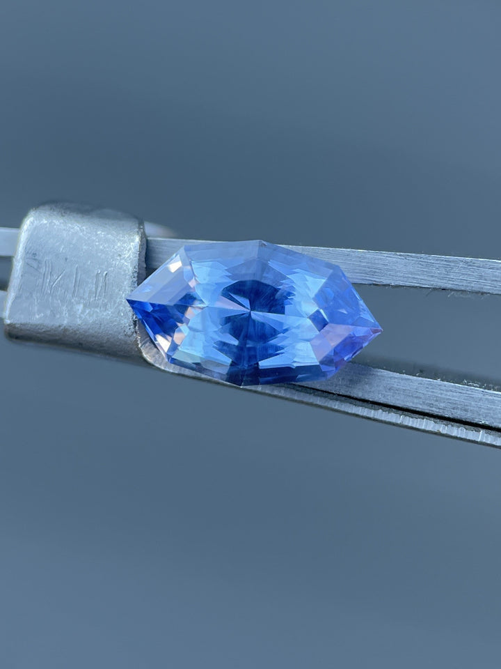 A stunning 1.73 carat sapphire held in tweezers, displaying vibrant denim blue and cornflower hues, cut in a custom geometric marquise shape, from Sri Lanka with no treatment.
