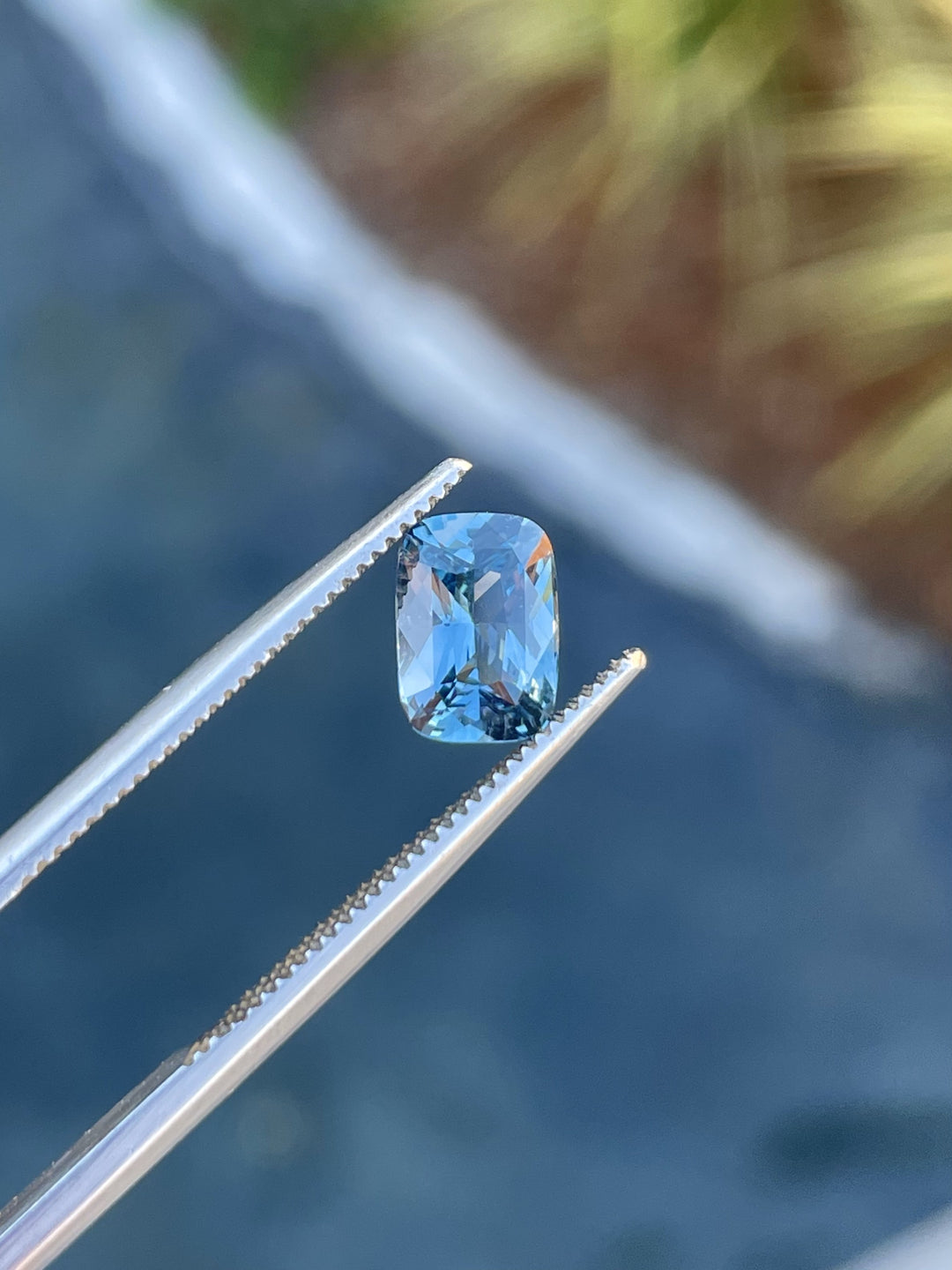 A 1.52-carat mix cushion-cut denim blue and green sapphire held in tweezers, showcasing its precision cut and clarity.