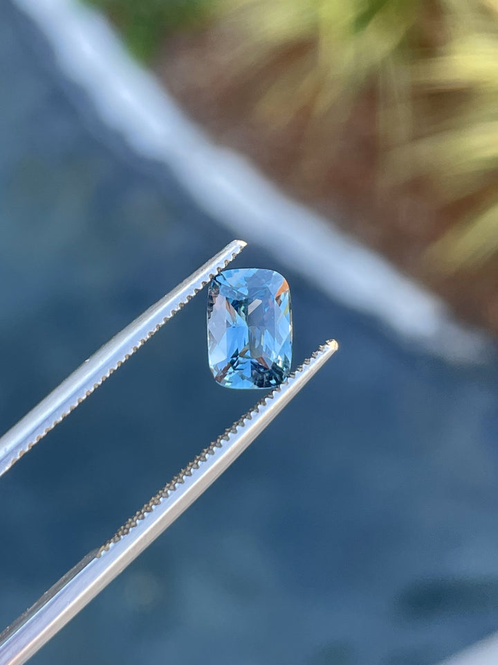 A 1.52-carat mix cushion-cut denim blue and green sapphire held in tweezers, showcasing its precision cut and clarity.
