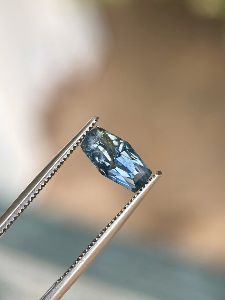 A 2.14-carat denim blue sapphire with a custom geometric elongated cushion cut, held by tweezers, revealing its precise cut and vibrant blue color. The gemstone from the Umba River, Tanzania, is untreated and measures 10.66mm by 5.55mm by 4.17mm.