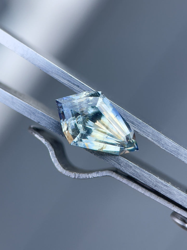 Close-up view of a 2.32 carat sapphire with yellow and Ceylon denim tones, displayed in a geometric step cut kite design, sourced from Sri Lanka, measuring 11.90 x 7.43 x 4.46 mm, held in metal clamps for inspection.