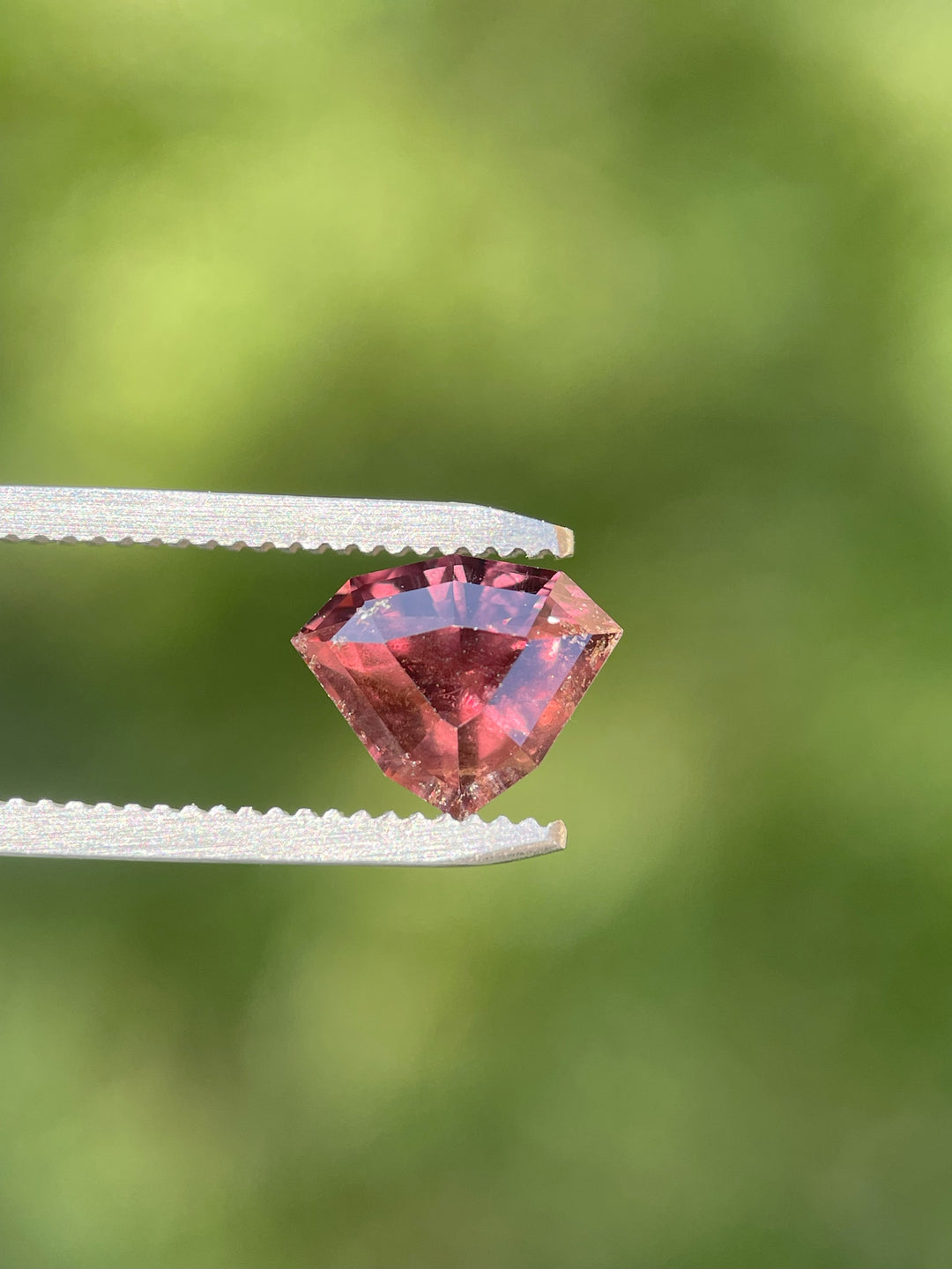 Side profile of a 2.94ct shield step-cut sapphire with deep strawberry red and mauve hues held by gem tweezers, highlighting the stone’s faceted depth and brilliance.