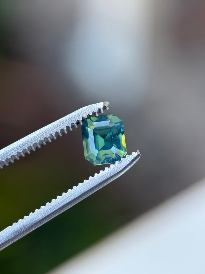 Green blue emerald cut opalescent sapphire held in gem tweezers showing off natural color and precision faceting