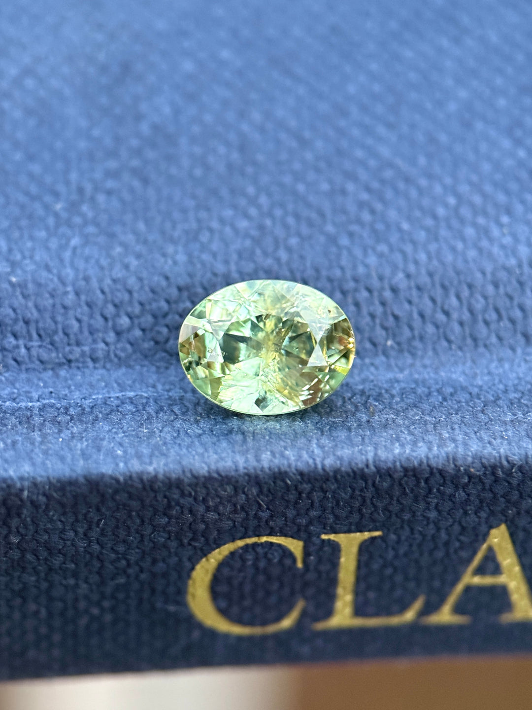 Earthy teal green Montana sapphire in a custom modified brilliant oval cut resting on a blue surface, showing yellowish undertones, natural color zoning, and crisp facet reflections in unheated sapphire.