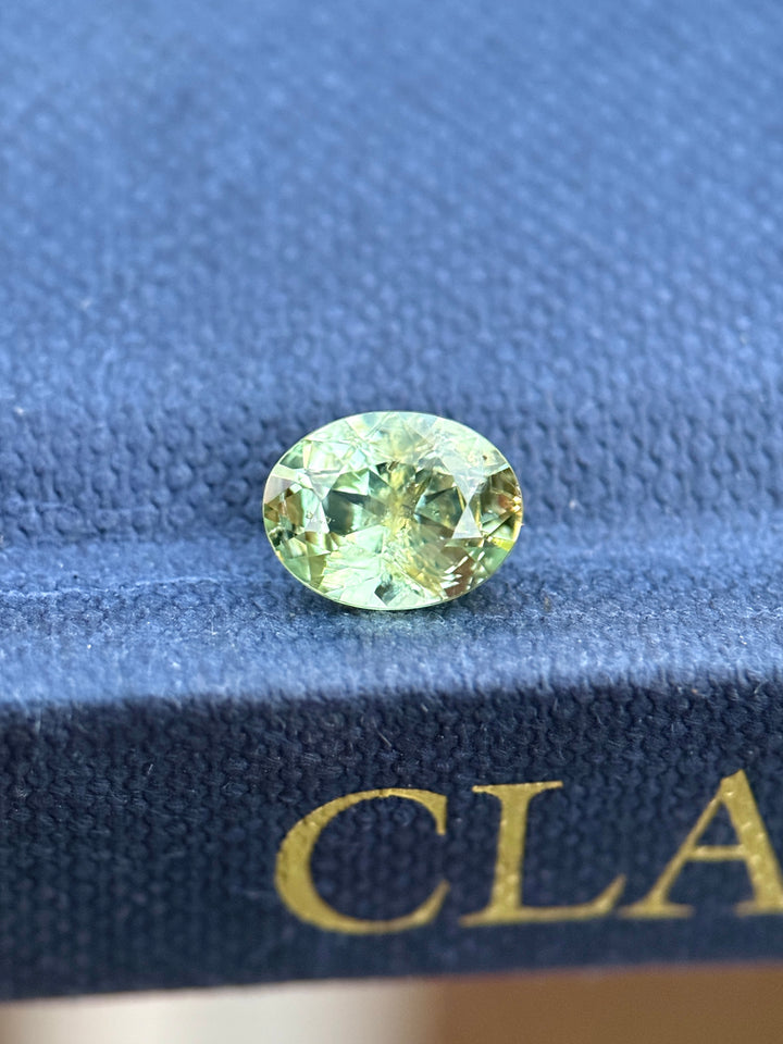 Earthy teal green Montana sapphire in a custom modified brilliant oval cut resting on a blue surface, showing yellowish undertones, natural color zoning, and crisp facet reflections in unheated sapphire.