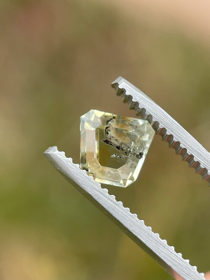 A 1.68-carat earthy yellow-green sapphire held in tweezers, showcasing its vibrant hues and shield tablet cut details against a natural setting.