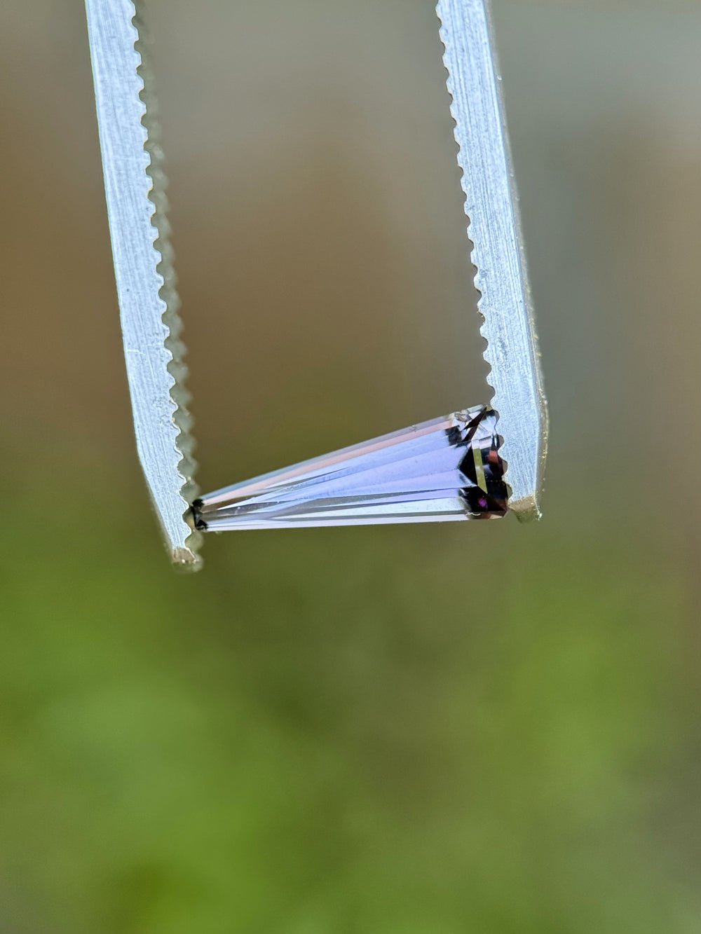 Close-up of a 0.67-carat Tanzanite held in tweezers, highlighting the step cut tapered baguette shape and color zoning.