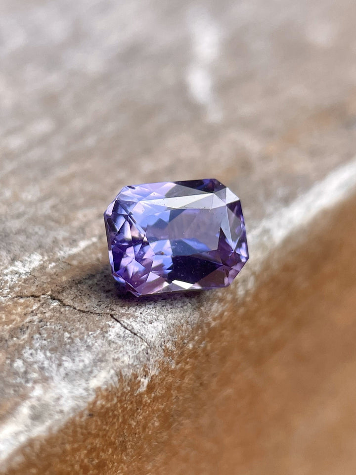 Close-up of a 1.05 carat pink and purple sapphire, radiant cut, resting on a textured wooden surface, showcasing its natural brilliance and origin from Sri Lanka.