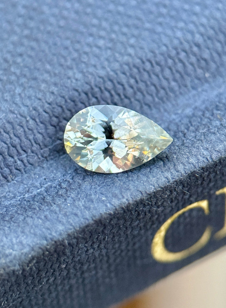 1.13 carat Montana sapphire in a precision pear mixed cut, photographed resting on a textured blue surface, showing faint silver seafoam tones and crisp natural brilliance in an untreated loose gemstone.