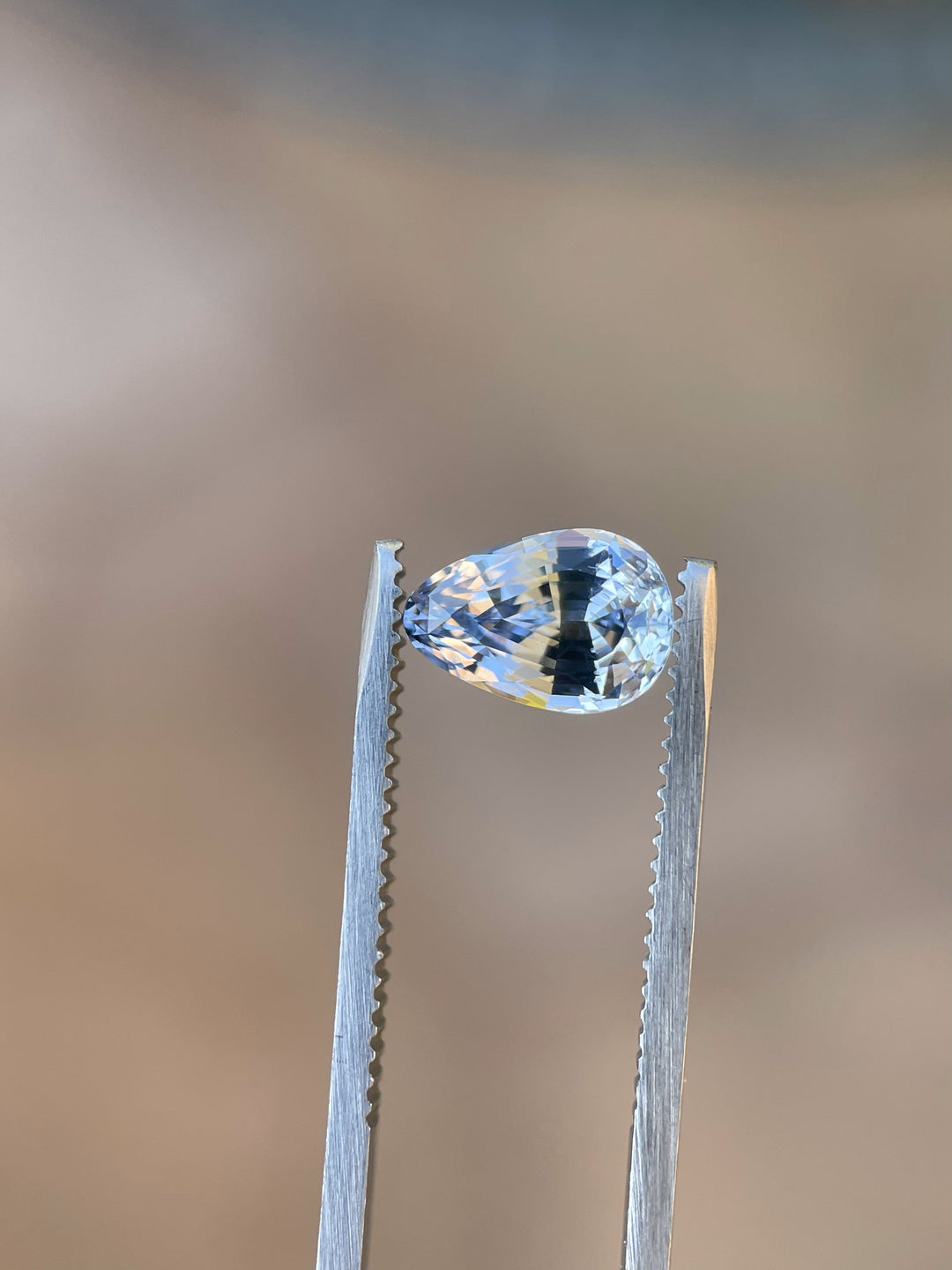 Close-up view of a tweezer holding a 2.39-carat cornflower and powder blue sapphire in a custom mix brilliant teardrop cut, emphasizing the gem's pristine clarity and vivid hues against a softly blurred background.