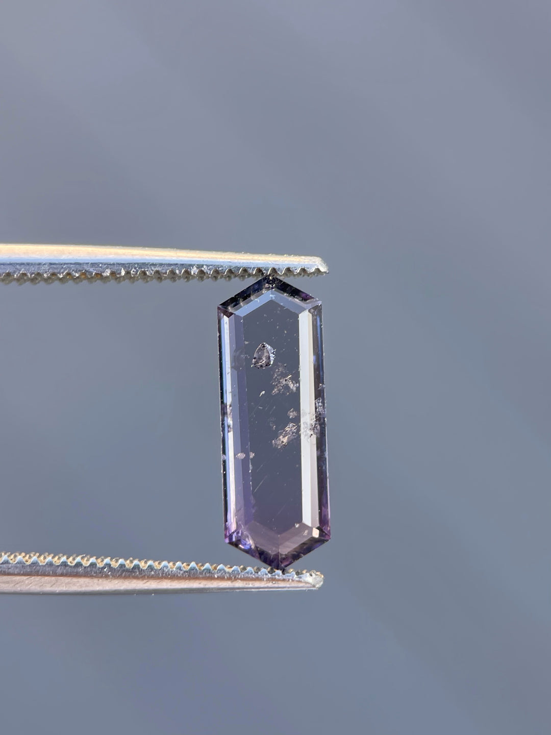 A purple and pink hexagonal step cut spinel held in precision tweezers, highlighting its elongated shape and geometric precision.