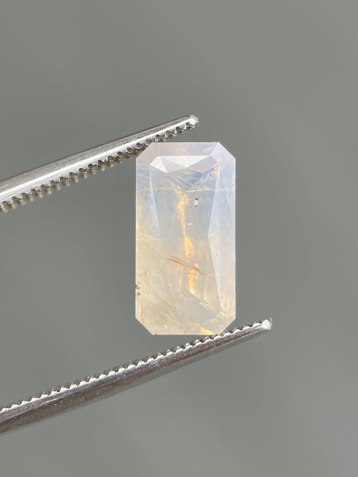 Close-up of a 4.18-carat opalescent white and yellow sapphire held by tweezers, showcasing its elongated radiant cut and natural inclusions.
