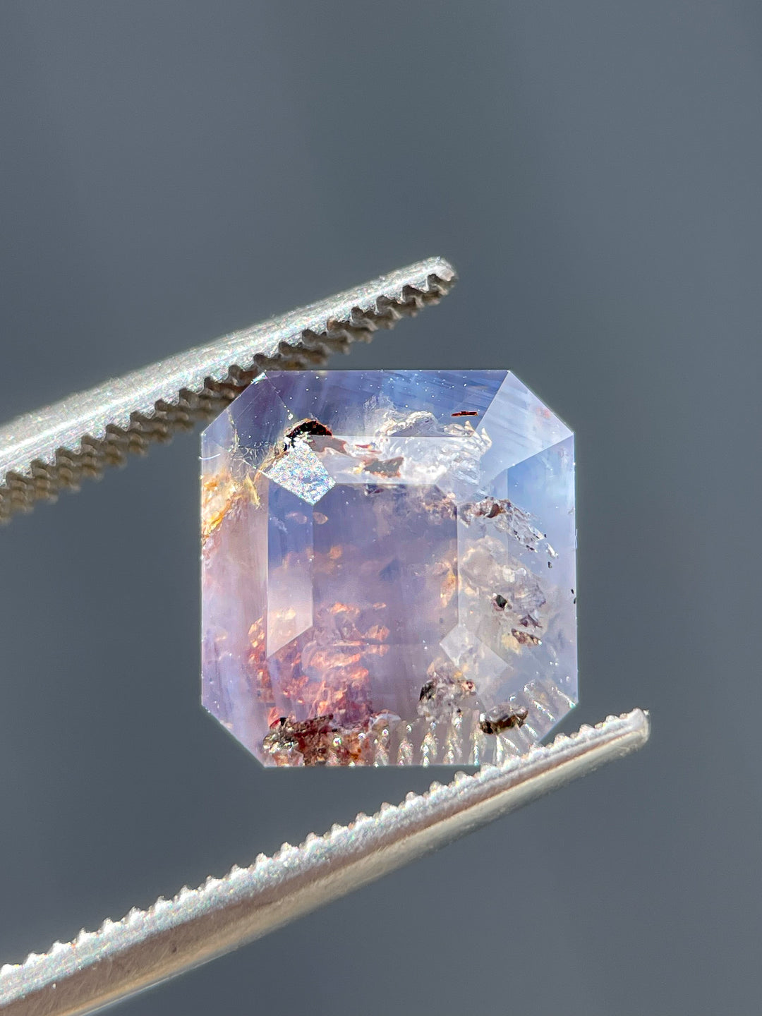 A stunning 5.92-carat emerald cut sapphire featuring gray, purple, and brown hues, with natural inclusions, expertly held in tweezers for detail.