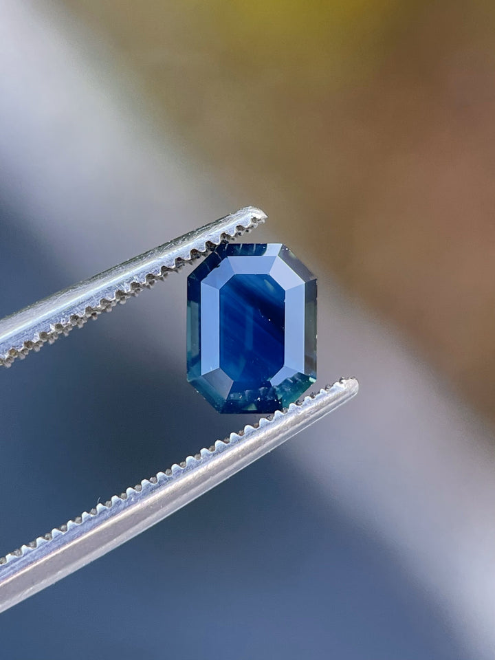 Close-up of a stunning teal-blue sapphire, held in tweezers to highlight its flawless octagonal step cut and vibrant color shift.