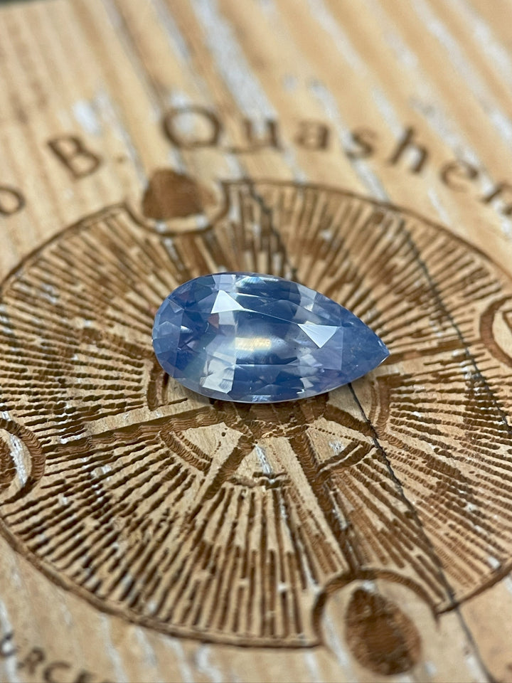 Close-up of an ethically sourced opalescent blue sapphire, featuring a mix brilliant pear cut and weighing 3.53 carats, placed on a decorative surface with detailed engraving.