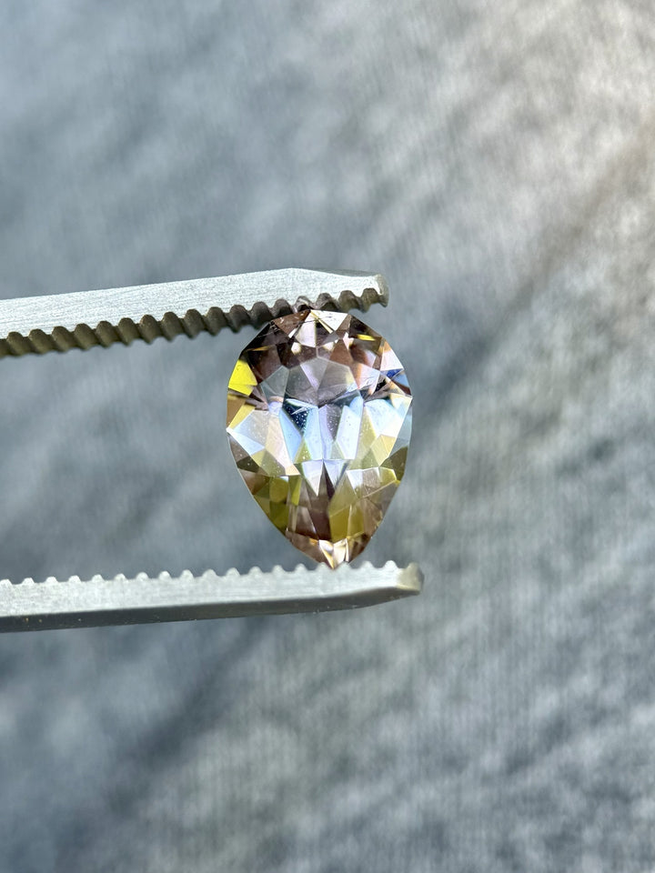 Close-up of custom pear-cut Tanzanite held by gem tweezers, showing vivid peach, pink, and purple color shifts