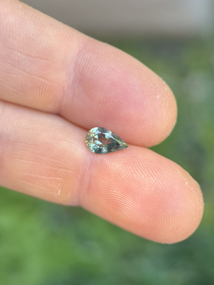 Loose 1.06ct sage green sapphire in modified pear brilliant cut displayed on fingertip, ethically sourced from Umba River, Tanzania