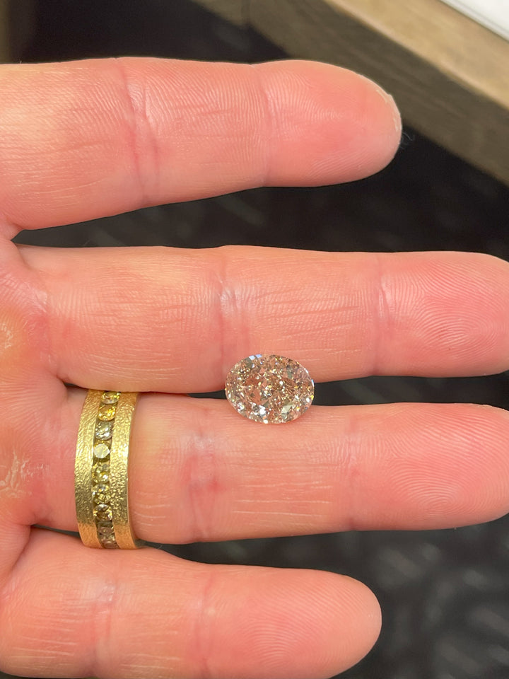 Hand holding a 5.25-carat oval champagne diamond, emphasizing its impressive size and light champagne color with strong rosiness. A gold ring with embedded yellow diamonds is visible in the background.