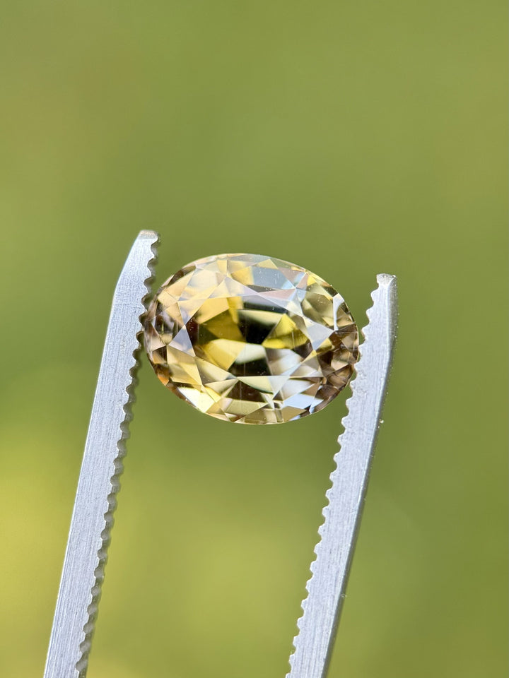 3.70 carat custom oval Tanzanite held in gemstone tweezers against soft green background, showing golden and peach flashes