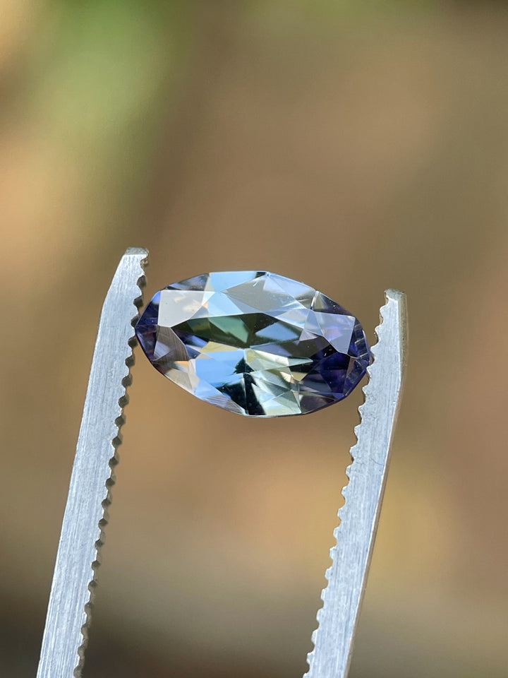 Teal and purple Tanzanite in precision oval cut held by tweezers, highlighting color shift and brilliance.