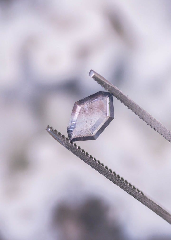 1.22 carat purple brown gray parti sapphire held in gem tweezers, highlighting geometric shield portrait cut facets and natural color zoning from Sri Lanka.