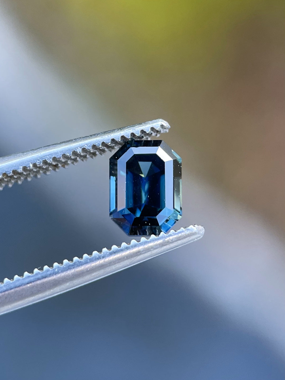 Close-up of a 1.16ct emerald-cut Nigerian sapphire held in precision tweezers, showing its deep green-blue hues and exquisite step-cut facets.