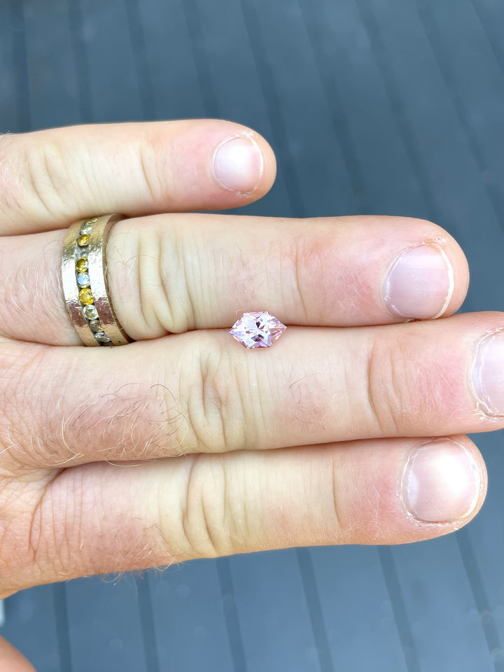 A 1.61 carat blush pink and peach sapphire held between fingers, showcasing its modified brilliant hexagon cut and vibrant color play. The natural light highlights the sapphire's unique brilliance and intricate facets.