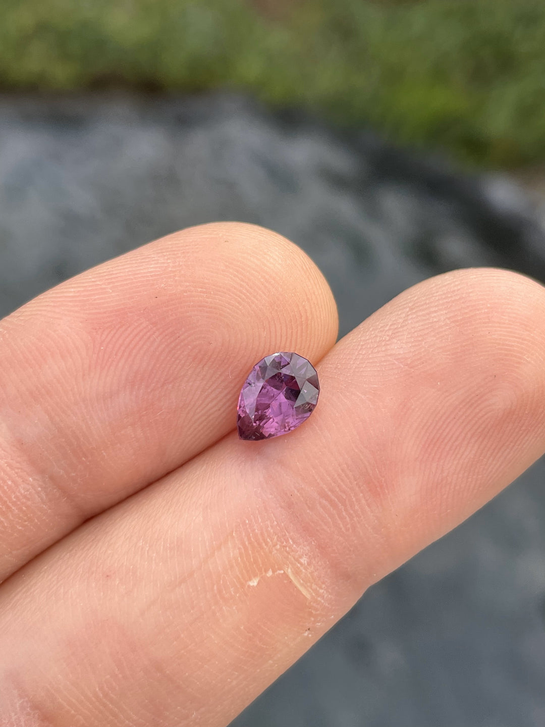 A close-up of a pinkish-purple pear-shaped sapphire placed on a person’s fingertip, showcasing its vibrant color and brilliance.