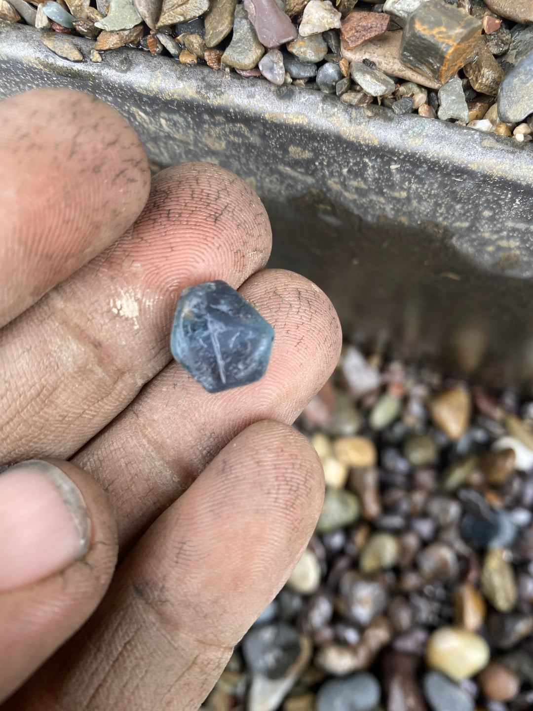 A close view of a hand holding a raw 5.79-carat hexagonal modified brilliant sapphire, freshly discovered and still dusty, set against a backdrop of assorted gravel, emphasizing the gem's natural origin from Montana, USA