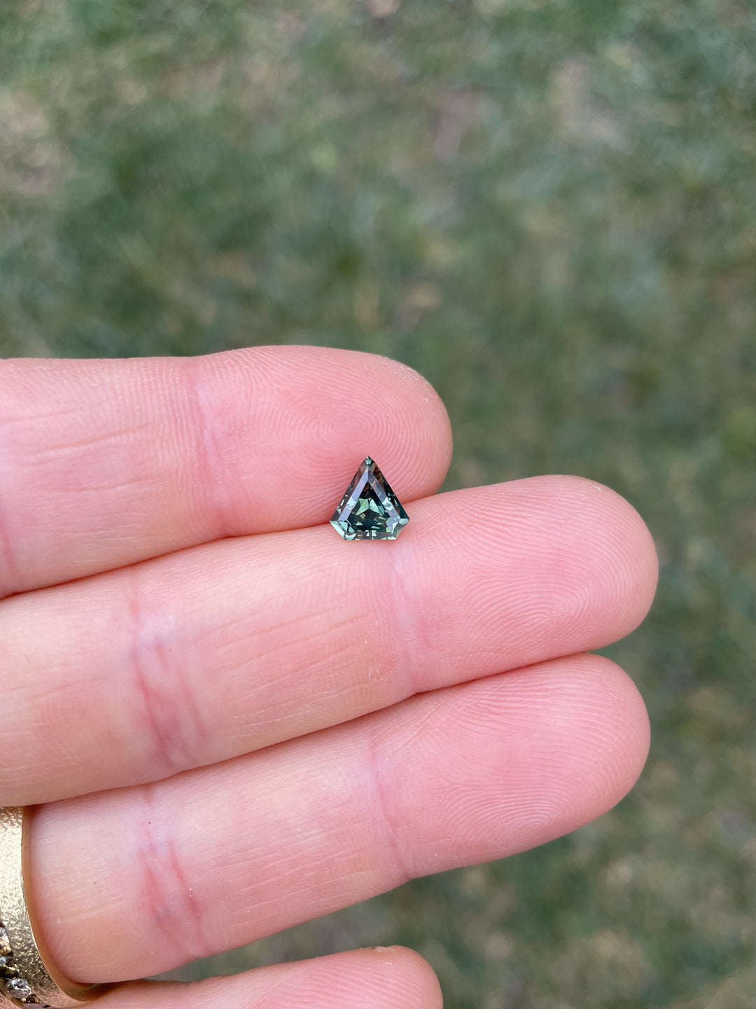 A 1.57ct sapphire with an opalescent yellow-teal and green color, shown on a fingertip to display its size and unique shield shape. The background is a blurred grassy field.