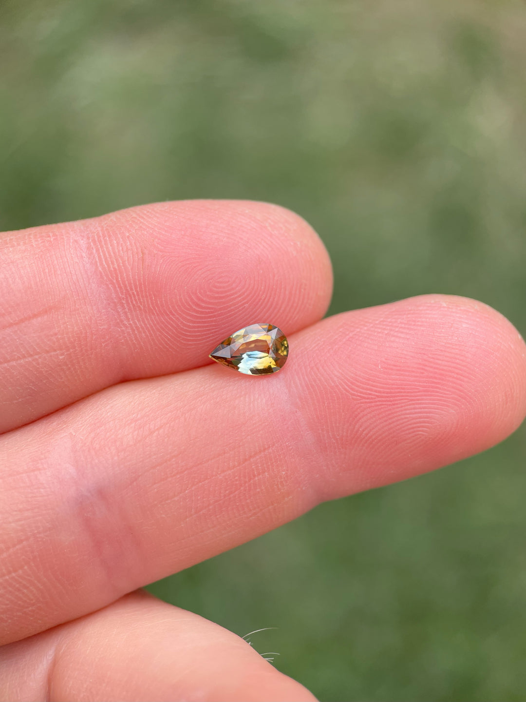 Close-up of a 1.00-carat bi-color pear sapphire resting on fingers, showcasing its brilliant mix of earthy brown and greenish-blue hues.