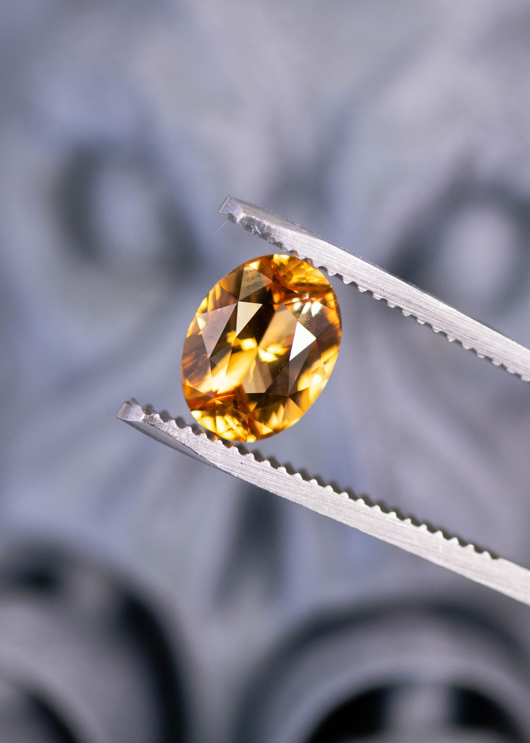 Heated orange sapphire oval gemstone held in tweezers, highlighting vivid yellow and red flashes and precision mixed brilliant faceting from Ratnapura, Sri Lanka